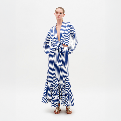 Against a plain white background, a person models the SVT DESIGNS Florina Skirt, featuring blue and white stripes and a tied waist. It includes long sleeves and a ruffled hemline. They also wear decorative sandals.