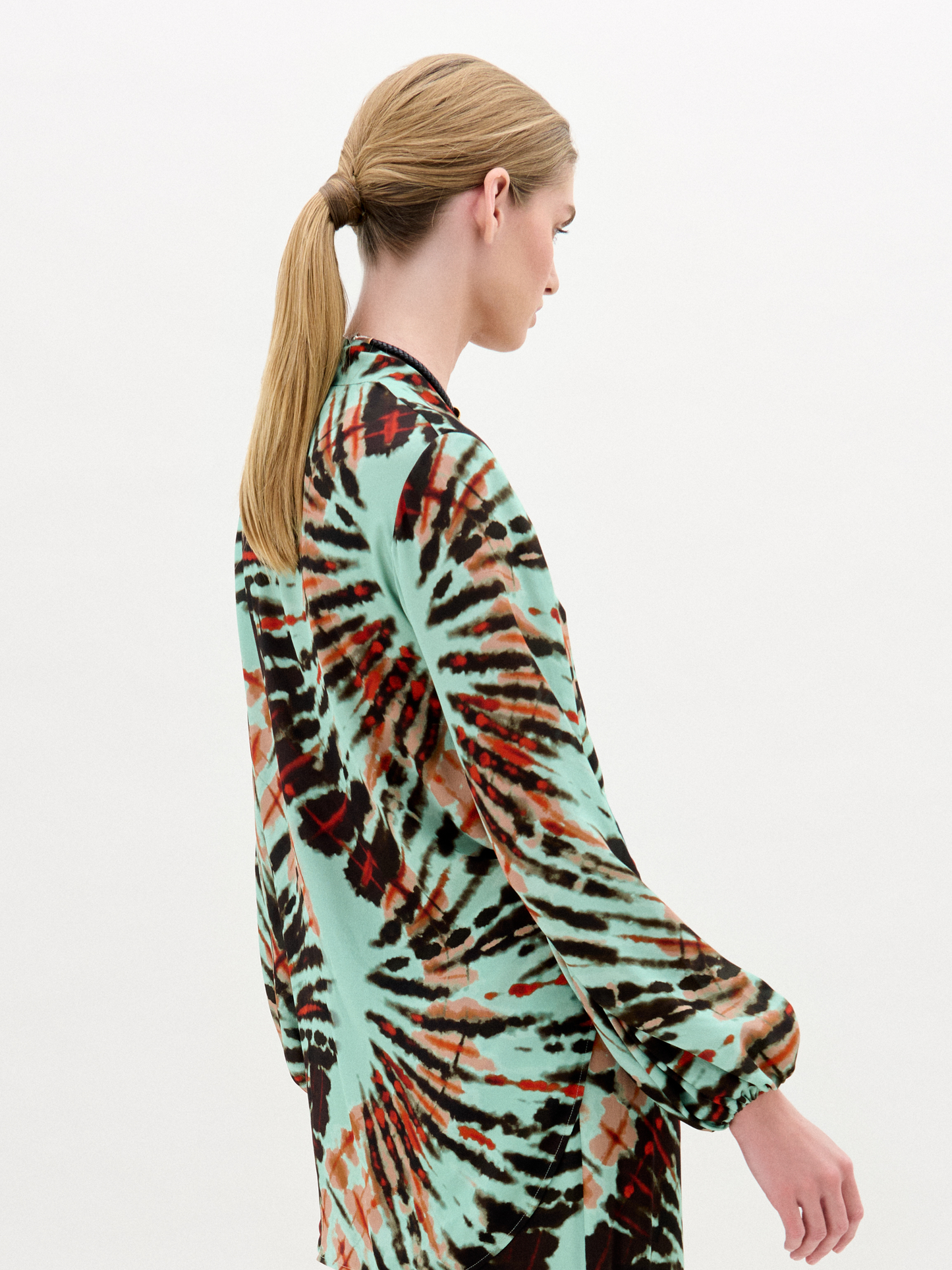 A woman stands against a plain white background wearing the Honey Blouse, a long-sleeved top featuring green, black, and orange abstract patterns with a chic front twist detail.
