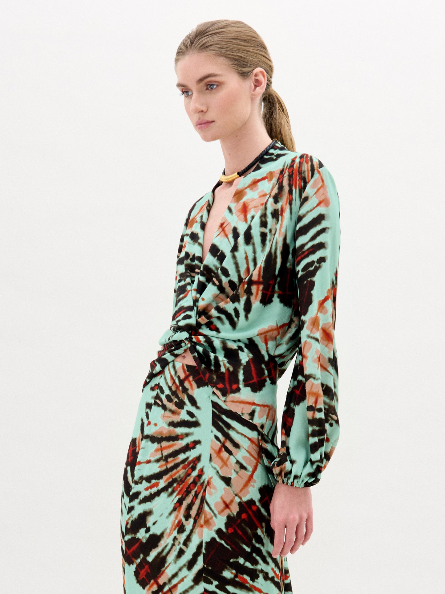 A woman with blonde hair in a low ponytail wears the SVT DESIGNS Honey Blouse, crafted from luxurious Italian fabric with a turquoise, black, and rust abstract pattern, standing against a plain white background.