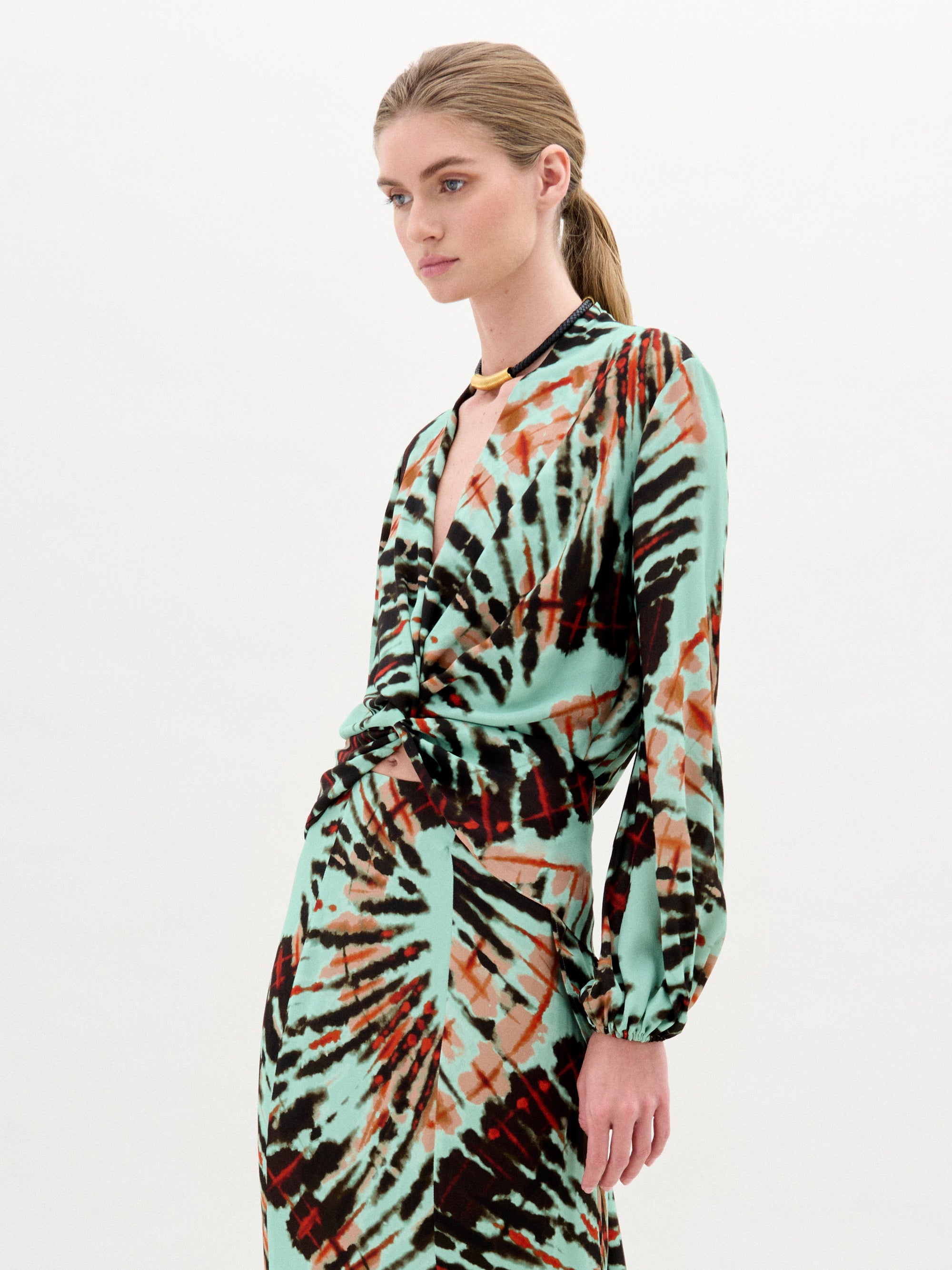 A woman with blonde hair in a low ponytail wears the SVT DESIGNS Honey Blouse, crafted from luxurious Italian fabric with a turquoise, black, and rust abstract pattern, standing against a plain white background.