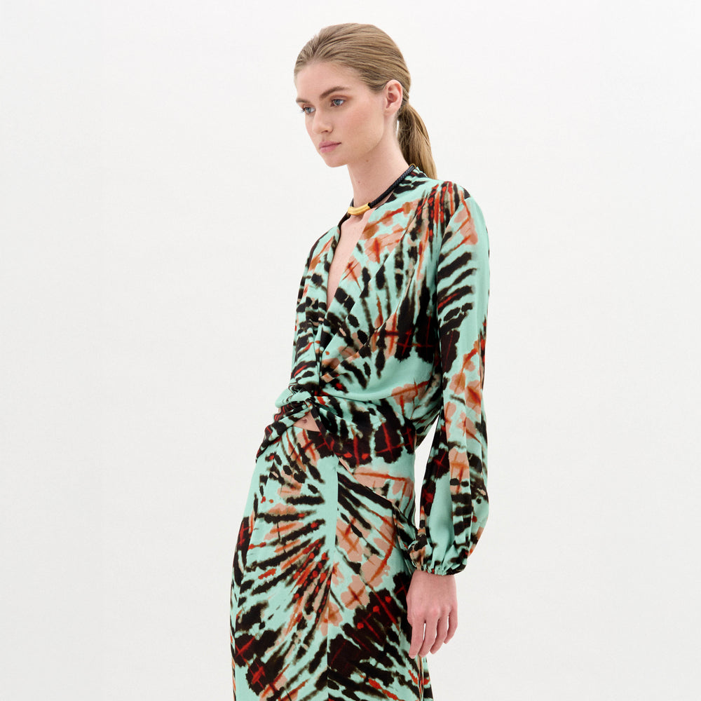 A person with light hair tied back is wearing the SVT DESIGNS Honey Blouse, a long-sleeved, tie-dye patterned top in mint green, black, and orange Italian fabric, standing against a plain white background.