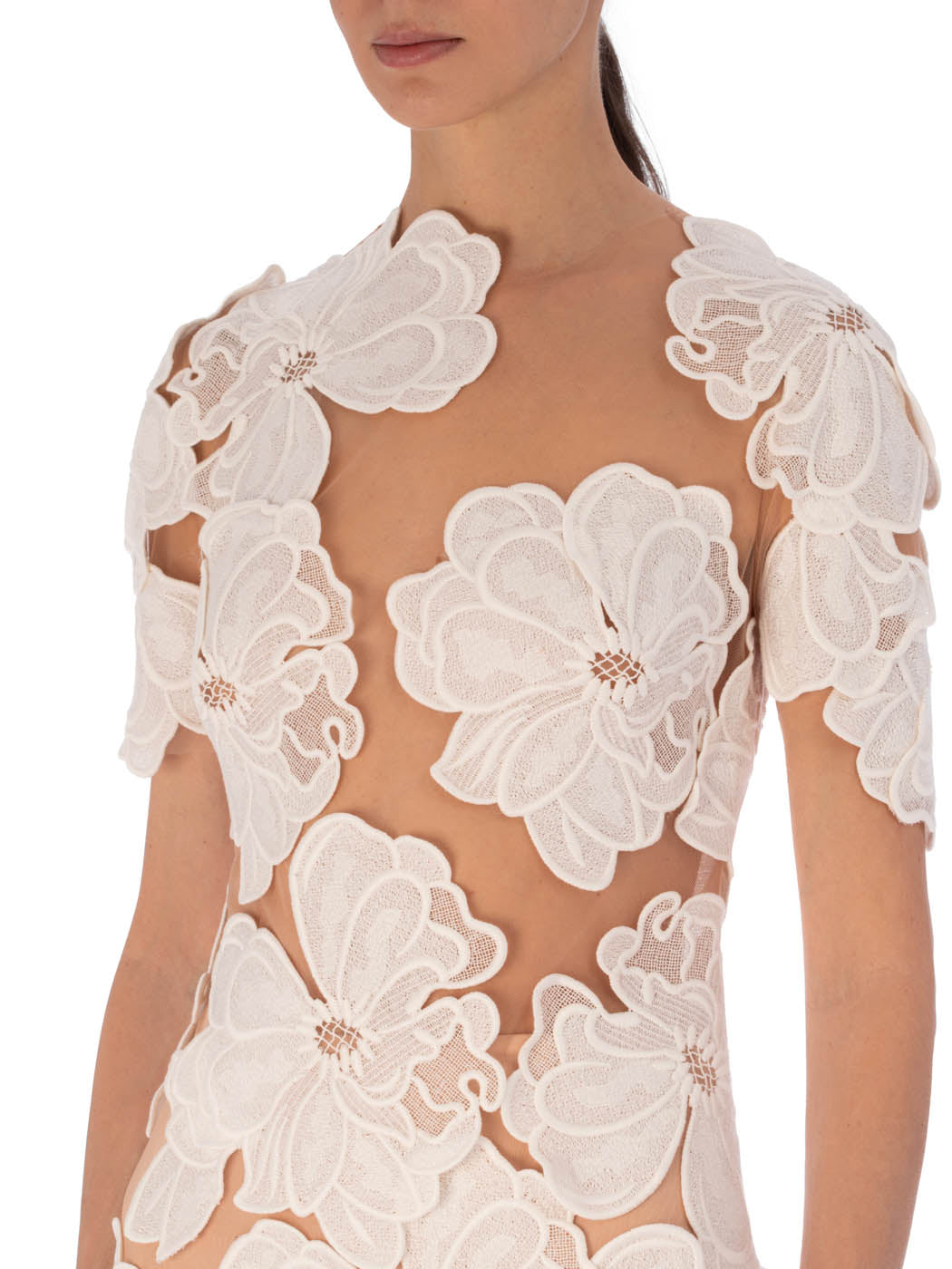 A woman wears the Hada Dress—a sheer mini dress with large white embroidered floral appliqués and floral lace—photographed from shoulders to waist against a plain background.