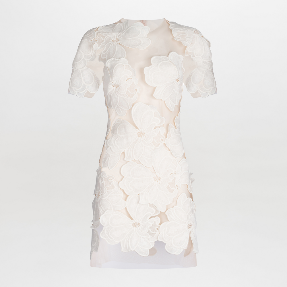 The Hada Dress is a short-sleeved nude mini dress featuring an asymmetric neckline and large white floral lace appliqué on the front for a textured, three-dimensional look. Shown against a plain white background.