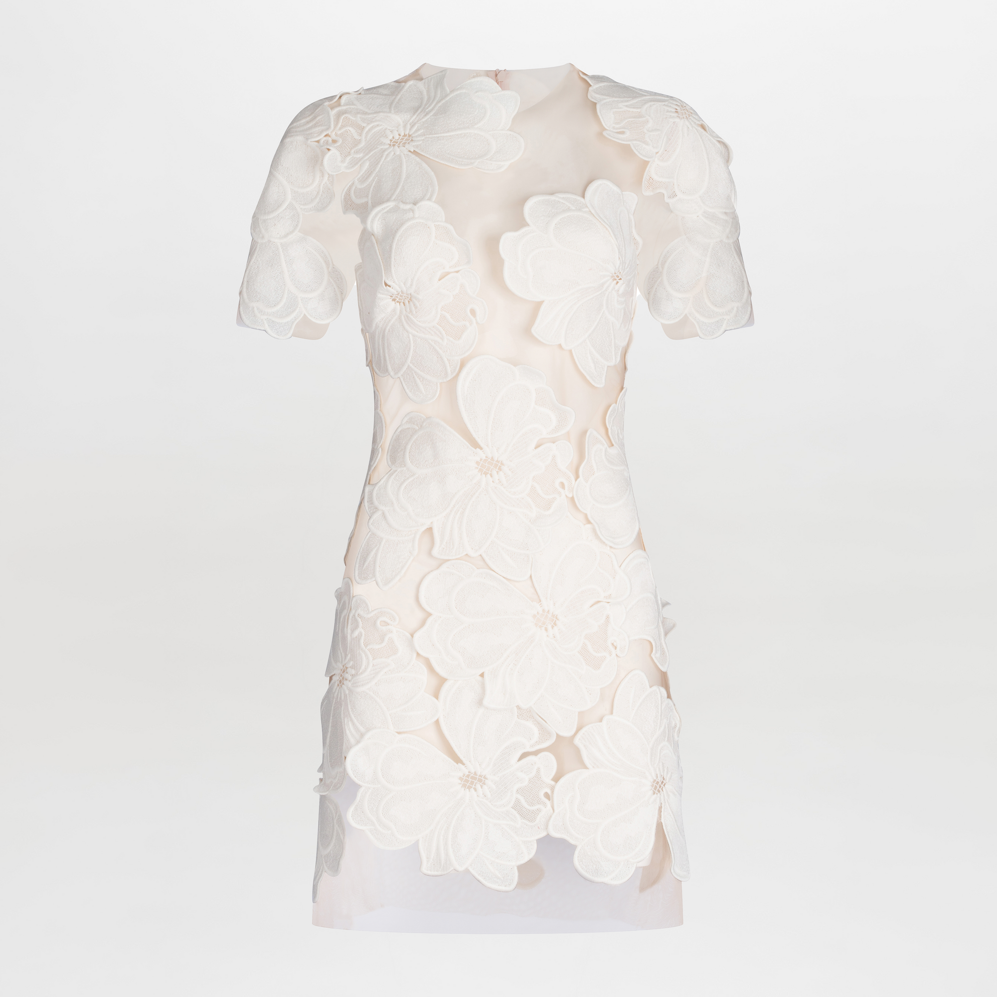 The Hada Dress is a short-sleeved nude mini dress featuring an asymmetric neckline and large white floral lace appliqué on the front for a textured, three-dimensional look. Shown against a plain white background.