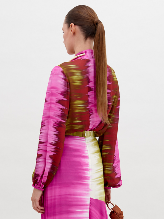 A woman with tied-back hair wears a Honey Blouse Violeta Lime Tie Dye with a front knot and matching skirt, accessorized with a gold bead necklace and holding a brown handbag against a plain background. This pre-order item will ship by November 15th, 2024.