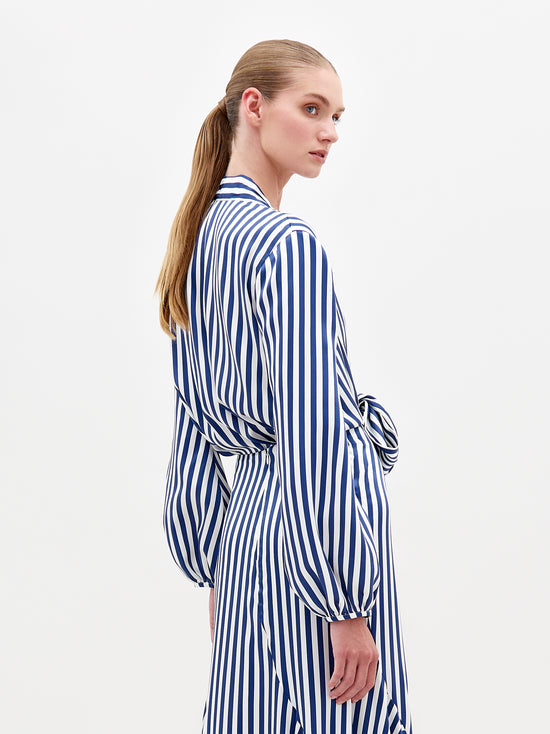 A person with long tied-back hair is wearing the Honey Blouse by SVT DESIGNS, featuring blue and white vertical stripes, a subtle plunging neckline, long sleeves, and a waist tie. The blouse is crafted from luxurious Italian textile. They stand sideways against a plain white background.