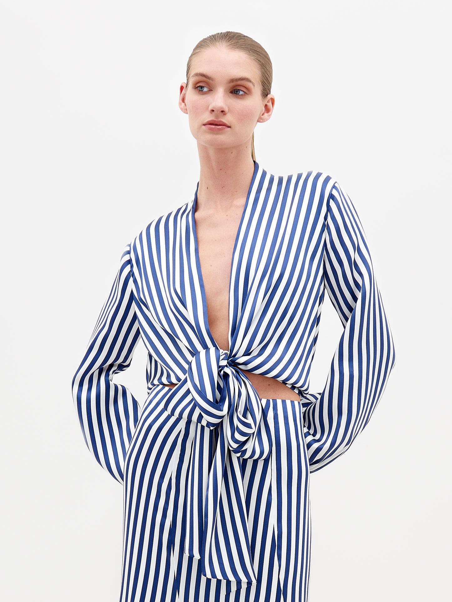 A person wears the SVT DESIGNS Honey Blouse, a blue and white vertically striped silk top with long sleeves and a tied front with a subtle plunging neckline. They stand against a plain background, showcasing effortless elegance from fine Italian textile while glancing to the side.