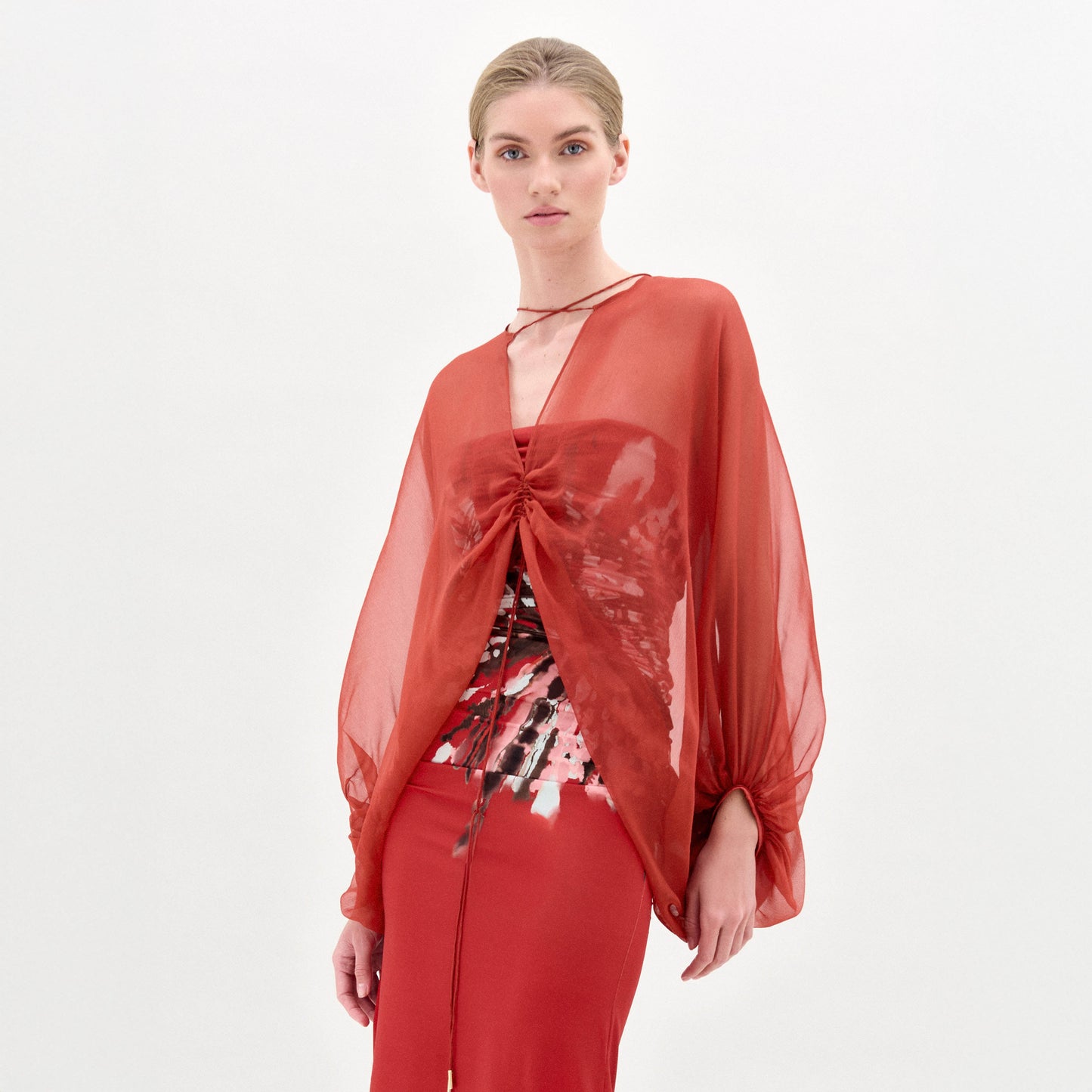 A woman stands against a plain background wearing the Idra Blouse, a sheer, long-sleeved top with a deep V neckline, layered over a patterned shirt and paired with a matching red skirt.