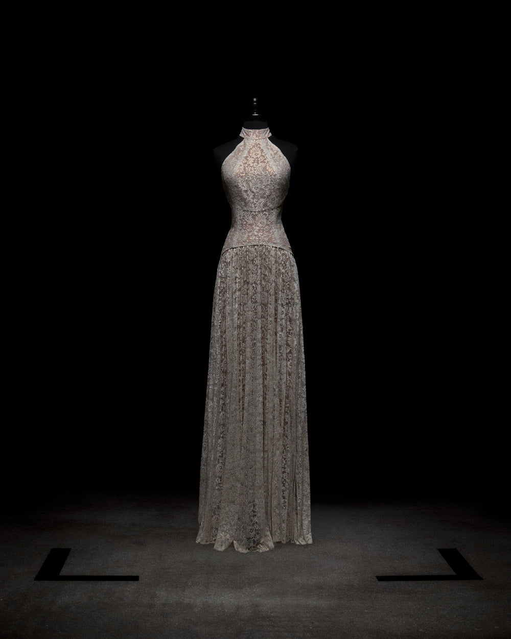The Ilana Dress by SVT DESIGNS is a sleeveless, floor-length evening gown with a high neck, showcased on a mannequin against a dark background. Crafted from delicate lace with intricate patterns, it exudes elegance.