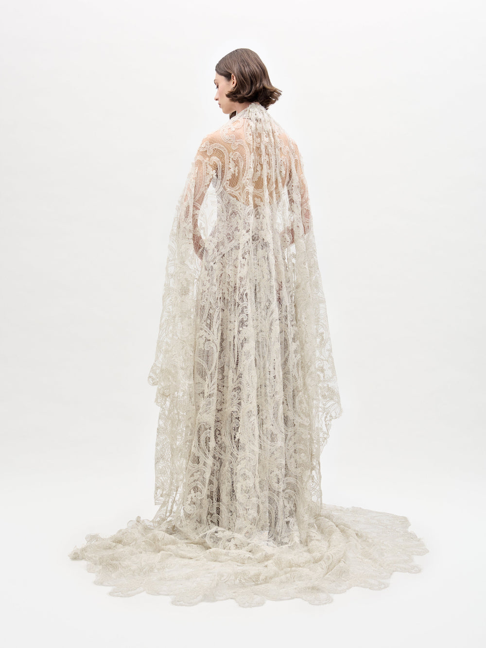 A person draped in the Olinta Wedding Veil by SVT DESIGNS stands back to camera against a white backdrop. The intricate lace patterns of the veil cascade onto the floor, creating an elegant trailing effect.