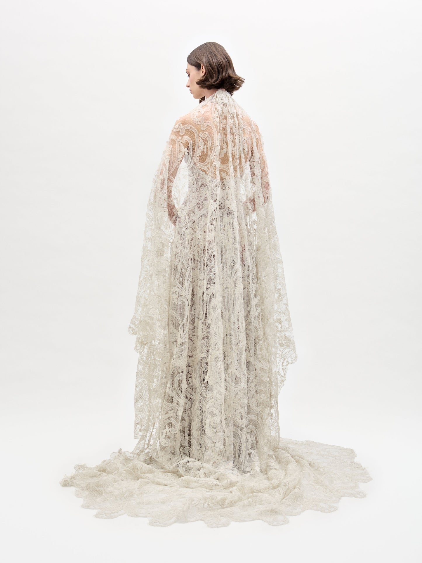 A person draped in the Olinta Wedding Veil by SVT DESIGNS stands back to camera against a white backdrop. The intricate lace patterns of the veil cascade onto the floor, creating an elegant trailing effect.