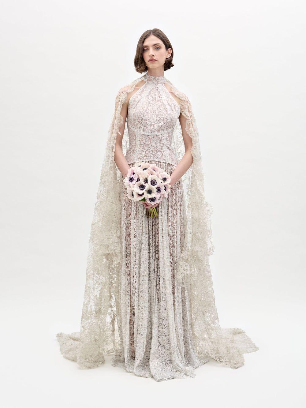 A woman models the SVT DESIGNS Olinta Wedding Veil against a plain background, wearing an intricate lace gown with a high neckline and cape. She holds a bouquet of white and purple flowers, and her hair is styled in a simple straight bob.