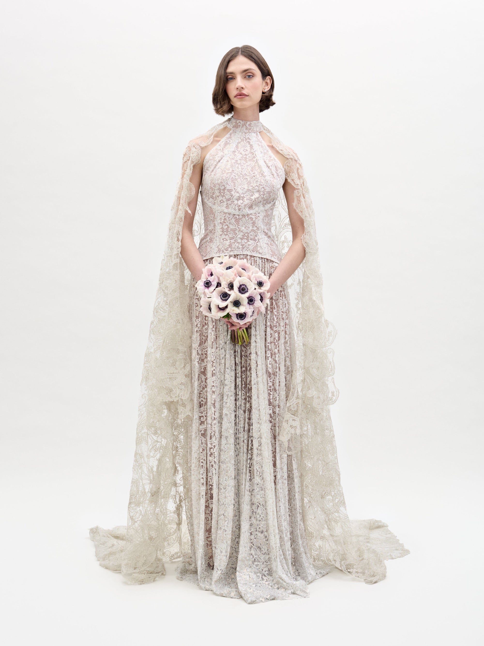 A woman models the SVT DESIGNS Olinta Wedding Veil against a plain background, wearing an intricate lace gown with a high neckline and cape. She holds a bouquet of white and purple flowers, and her hair is styled in a simple straight bob.