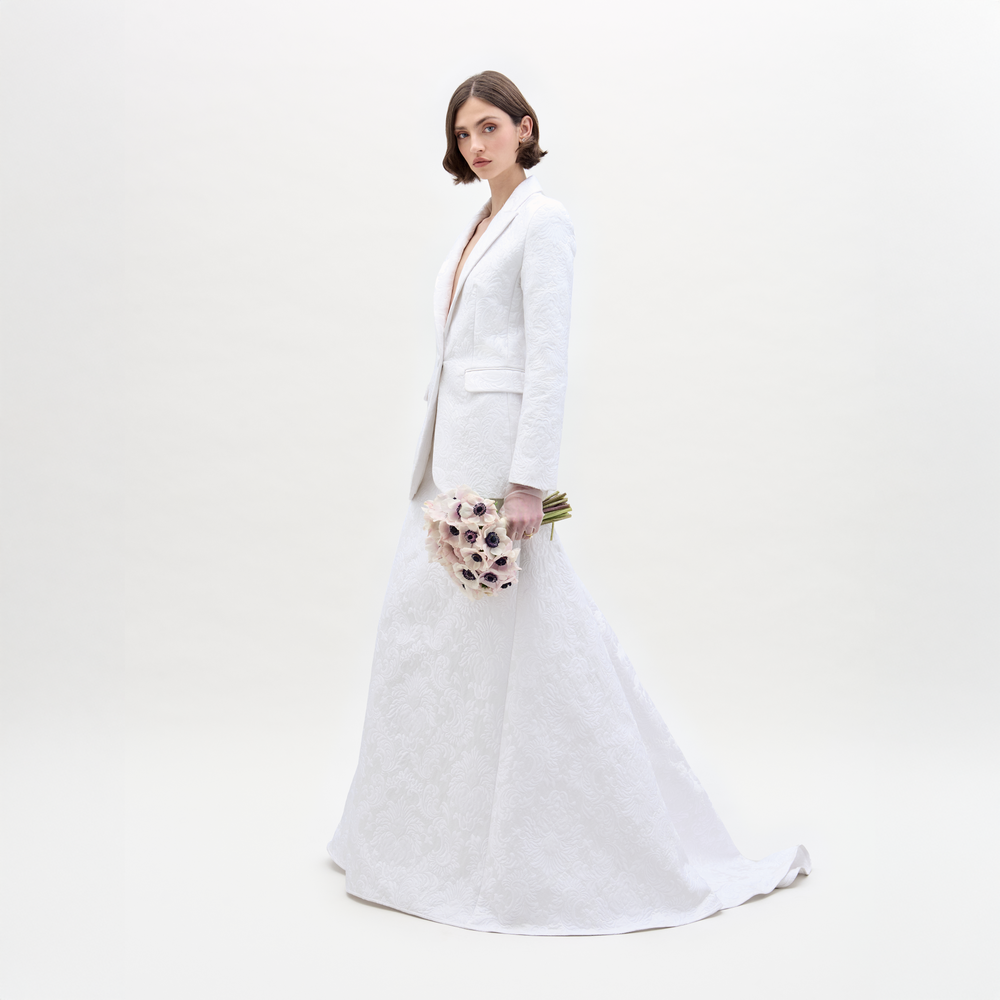 A woman wearing the SVT DESIGNS Imola Skirt and a white textured suit jacket stands against a plain white background, facing forward with a neutral expression and holding a bouquet of pale flowers with dark centers.
