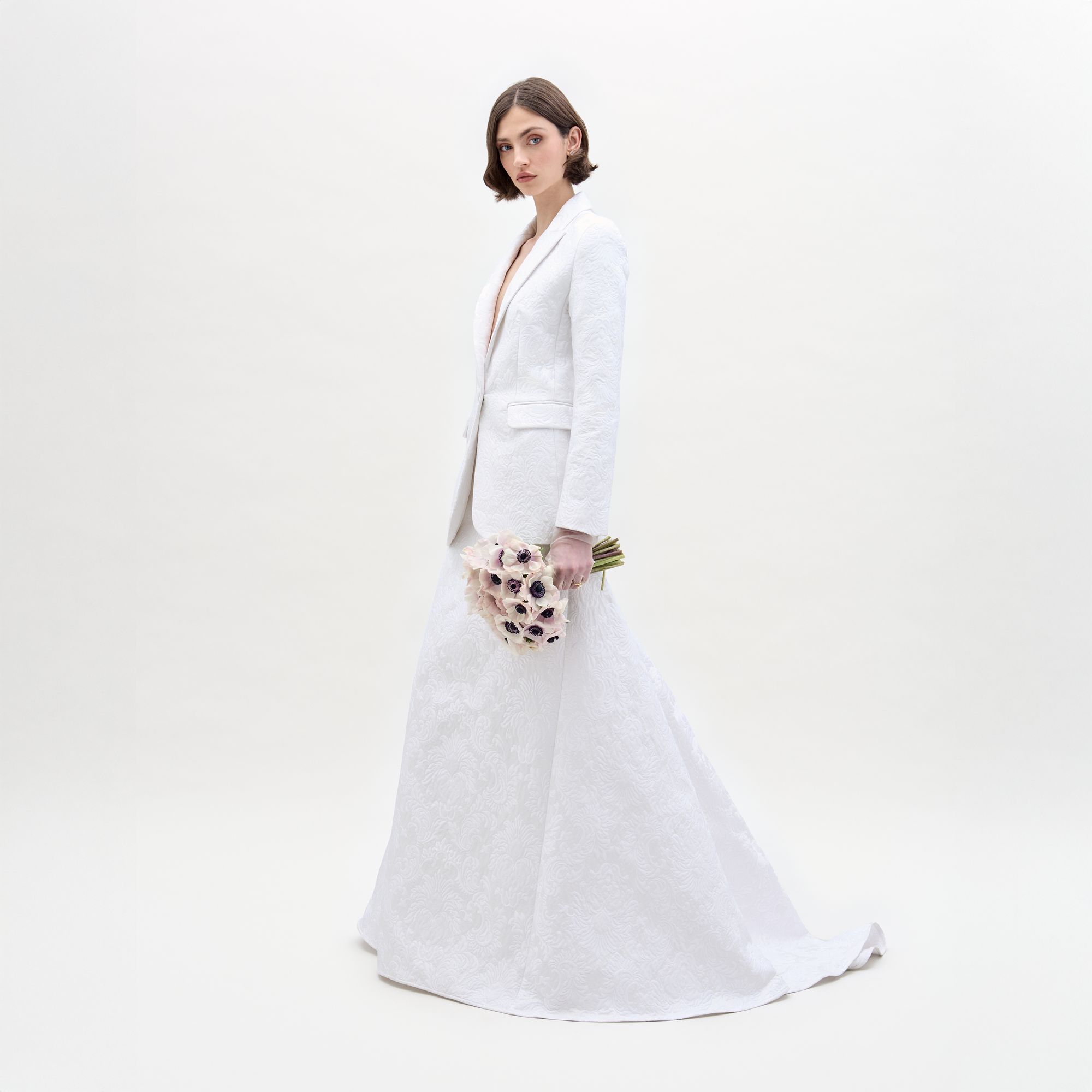 A woman wearing the SVT DESIGNS Imola Skirt and a white textured suit jacket stands against a plain white background, facing forward with a neutral expression and holding a bouquet of pale flowers with dark centers.
