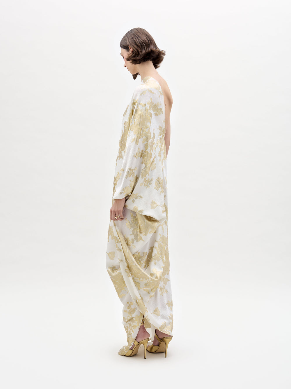A person with short brown hair stands in profile against a plain white background, wearing the elegant Inu Dress by SVT DESIGNS, featuring a loose, flowing white and gold floral design. Paired with gold high heels, they create a sophisticated look.