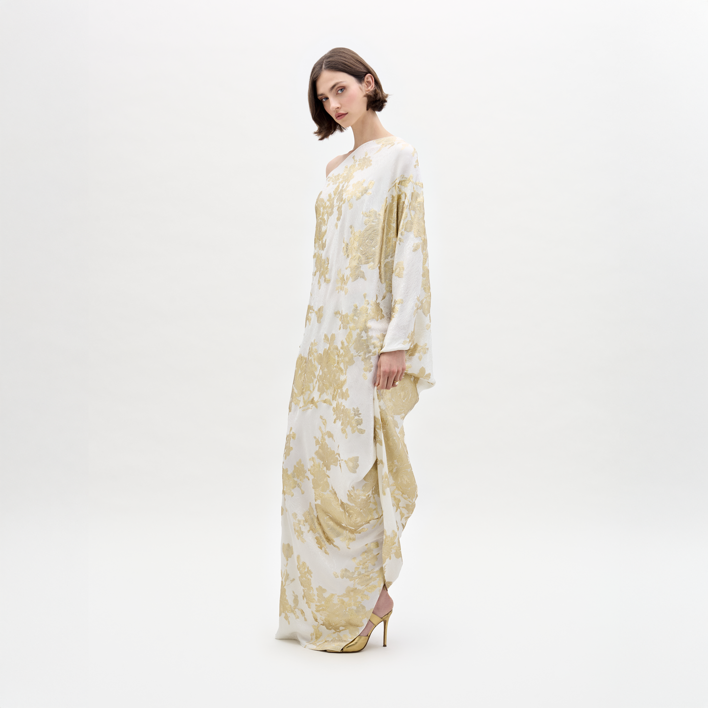 A woman with short dark hair gazes to the side, wearing the Inu Dress by SVT DESIGNS—a flowing silk maxi dress adorned with gold floral patterns—paired with high-heeled shoes against a plain white background.