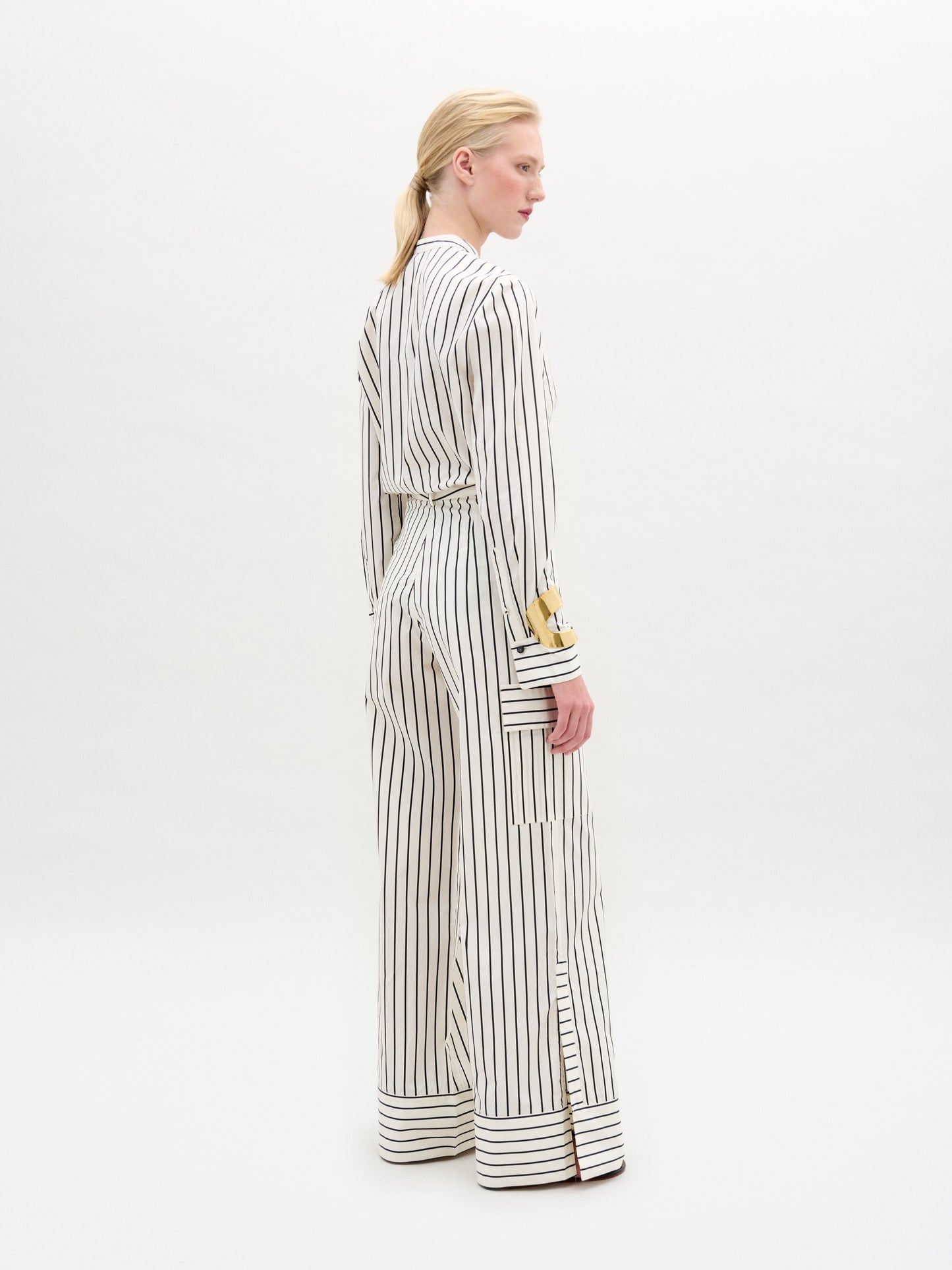 A person with long blonde hair stands sideways in a SVT DESIGNS white jumpsuit featuring black vertical stripes and wide-leg trousers, accented with gold buttons on sleeves and pockets, made from luxurious Italian fabric. The product is the Falak Pant, set against a plain white background.