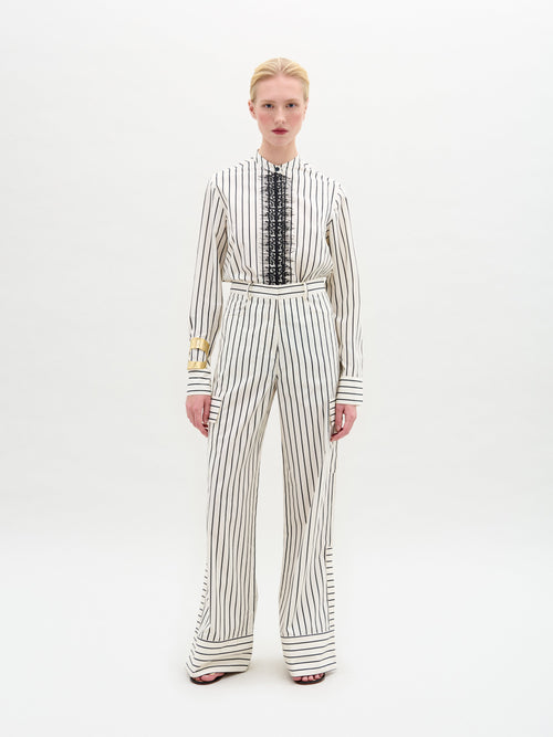 Dressed in SVT DESIGNS, a person models the Falak Pant, crafted from luxurious Italian fabric. The white outfit has black vertical stripes; the shirt features black lace details, and the high-waisted trousers are wide-leg. They maintain a neutral expression against a plain background.