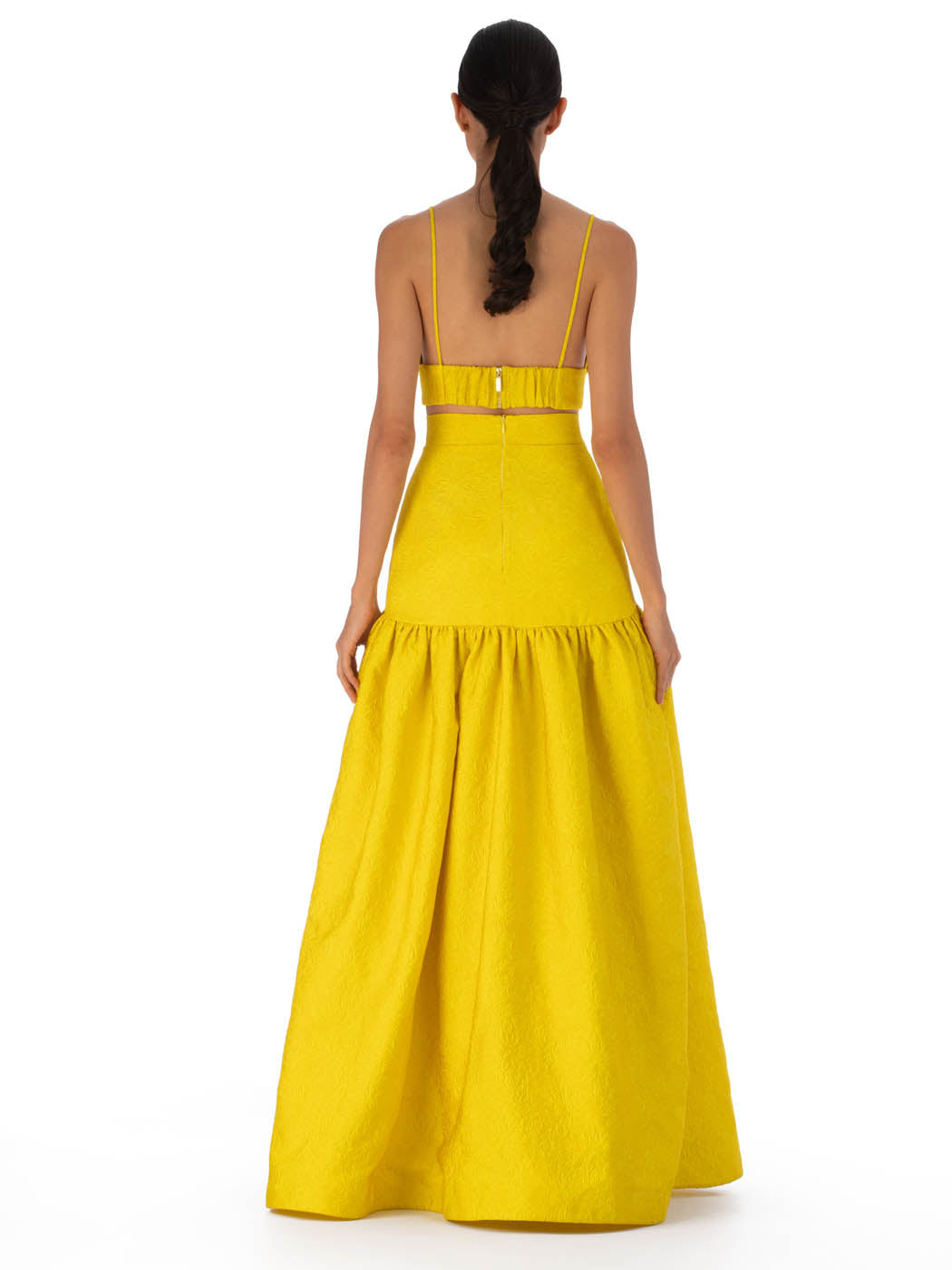 A woman with long dark hair stands away, wearing the SVT DESIGNS Ibbie Top. The top features a 3D floral design, thin straps, and a fitted style. She pairs it with a bright yellow floor-length skirt that flares from the waist. The background is plain white.