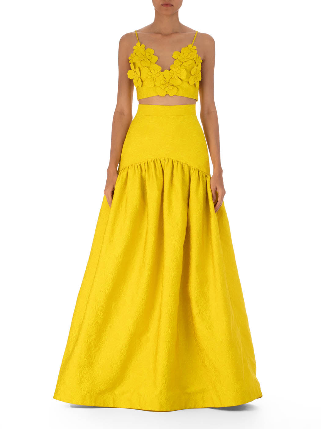 A person dons the Ibbie Top from SVT DESIGNS, a bright yellow sleeveless top with 3D floral designs, paired with a long voluminous skirt. The jacquard texture of the ensemble is accented against a plain white background.