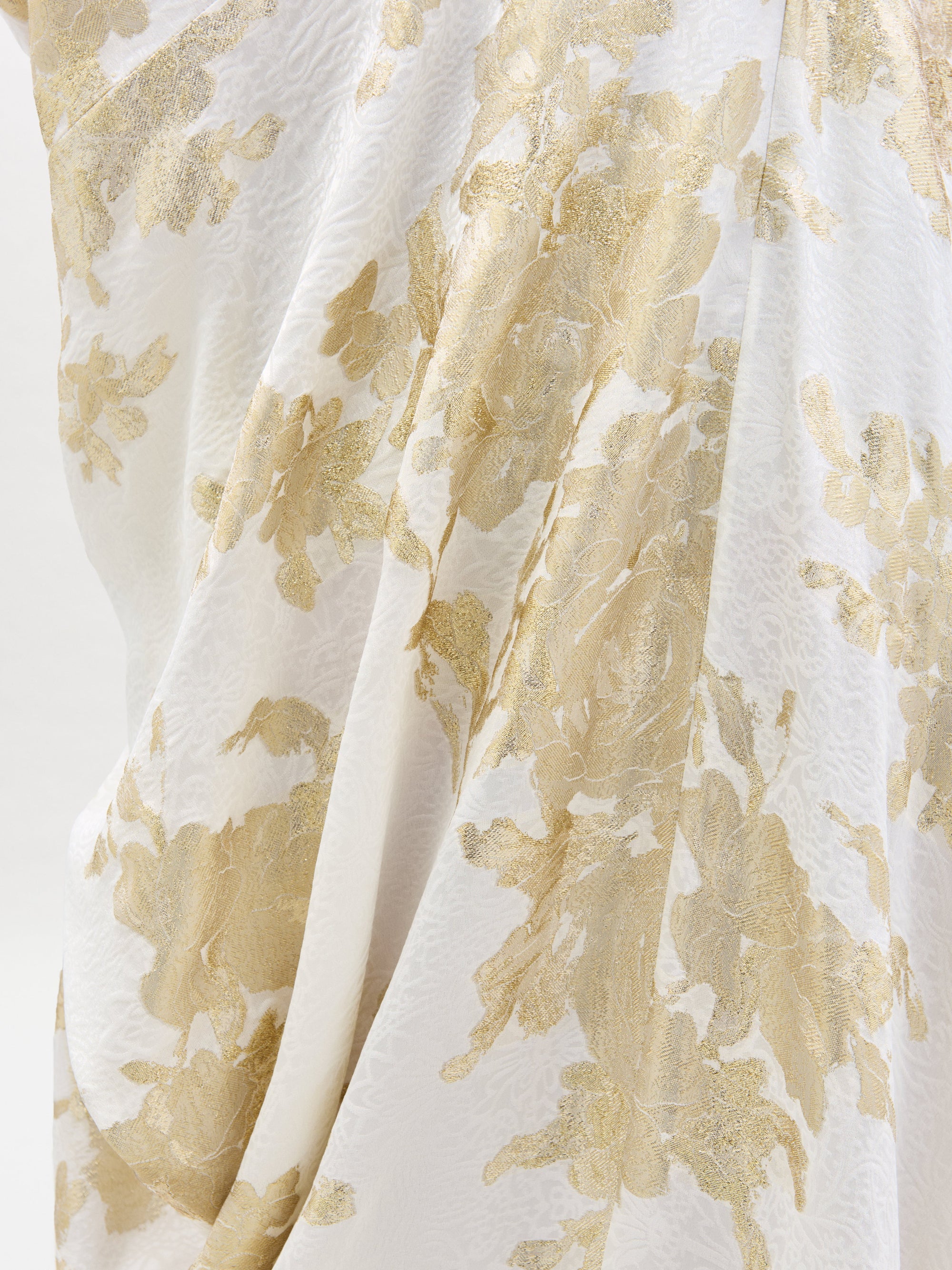The Inu Dress by SVT DESIGNS features fabric with a white base and an intricate metallic gold floral pattern, beautifully draped in soft folds. The textured, lustrous finish adds luxury and sophistication.