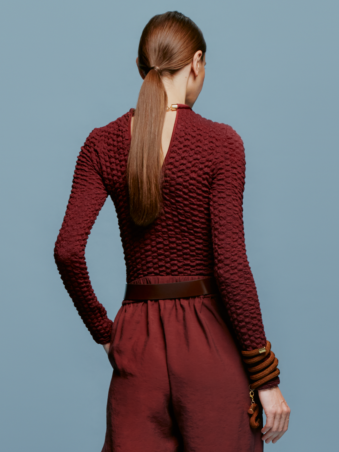 A person with long hair is wearing the Jari Top Burgundy, matching pants, and a wide belt, standing against a blue background like they're ready to set sail on a ship.