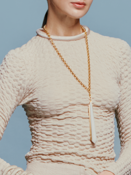 A woman with tied-back hair wears the Jari Top Cream, a long gold necklace, and several bracelets, standing against a light blue background reminiscent of the sky on August 19th.