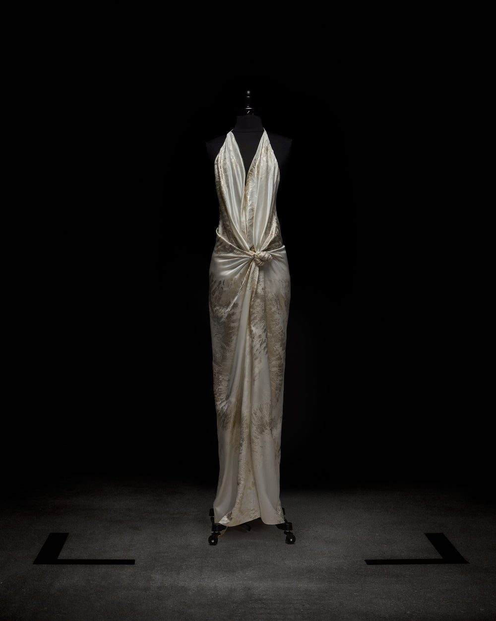 The Jazmin Dress by SVT DESIGNS, an ivory satin floor-length gown with a tie halter neckline and gathered knotted waist detail, is shown on a black dress form set against a dark background.