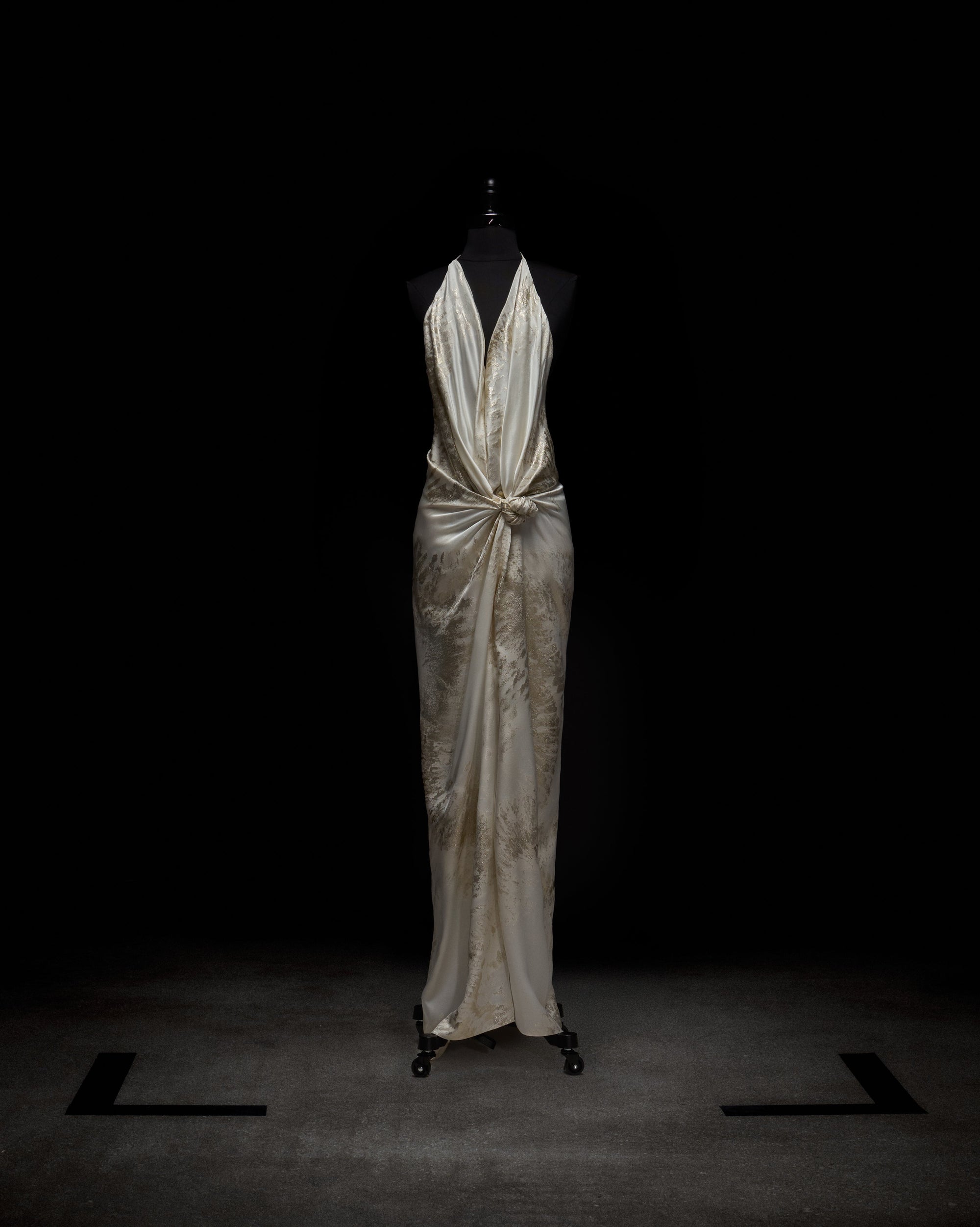 The Jazmin Dress by SVT DESIGNS, an ivory satin floor-length gown with a tie halter neckline and gathered knotted waist detail, is shown on a black dress form set against a dark background.