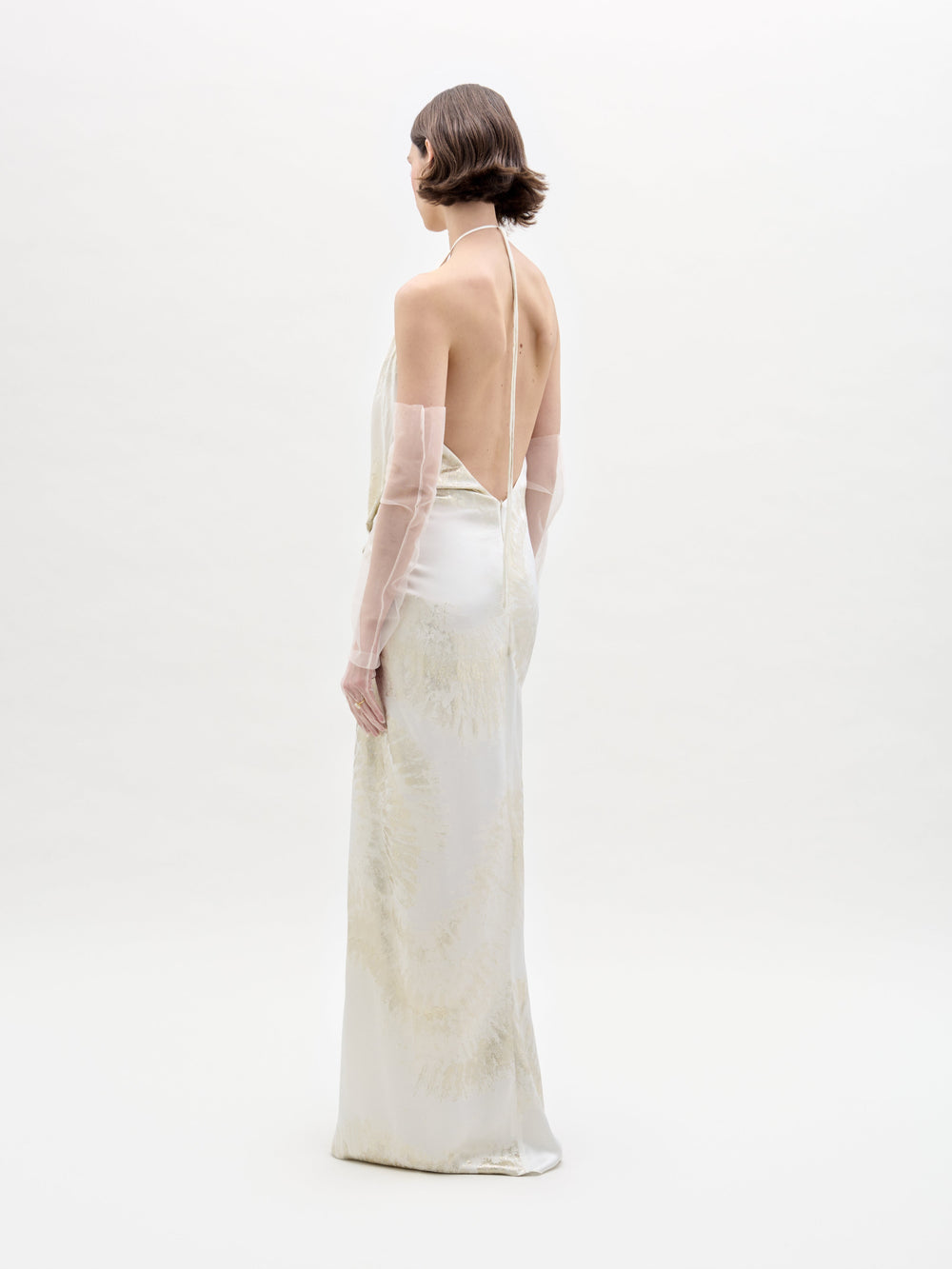A person with short brown hair, wearing the SVT DESIGNS Jazmin Dress—a long, backless white dress with a subtle pattern—and transparent elbow-length gloves, stands facing slightly to the side against a plain white background.