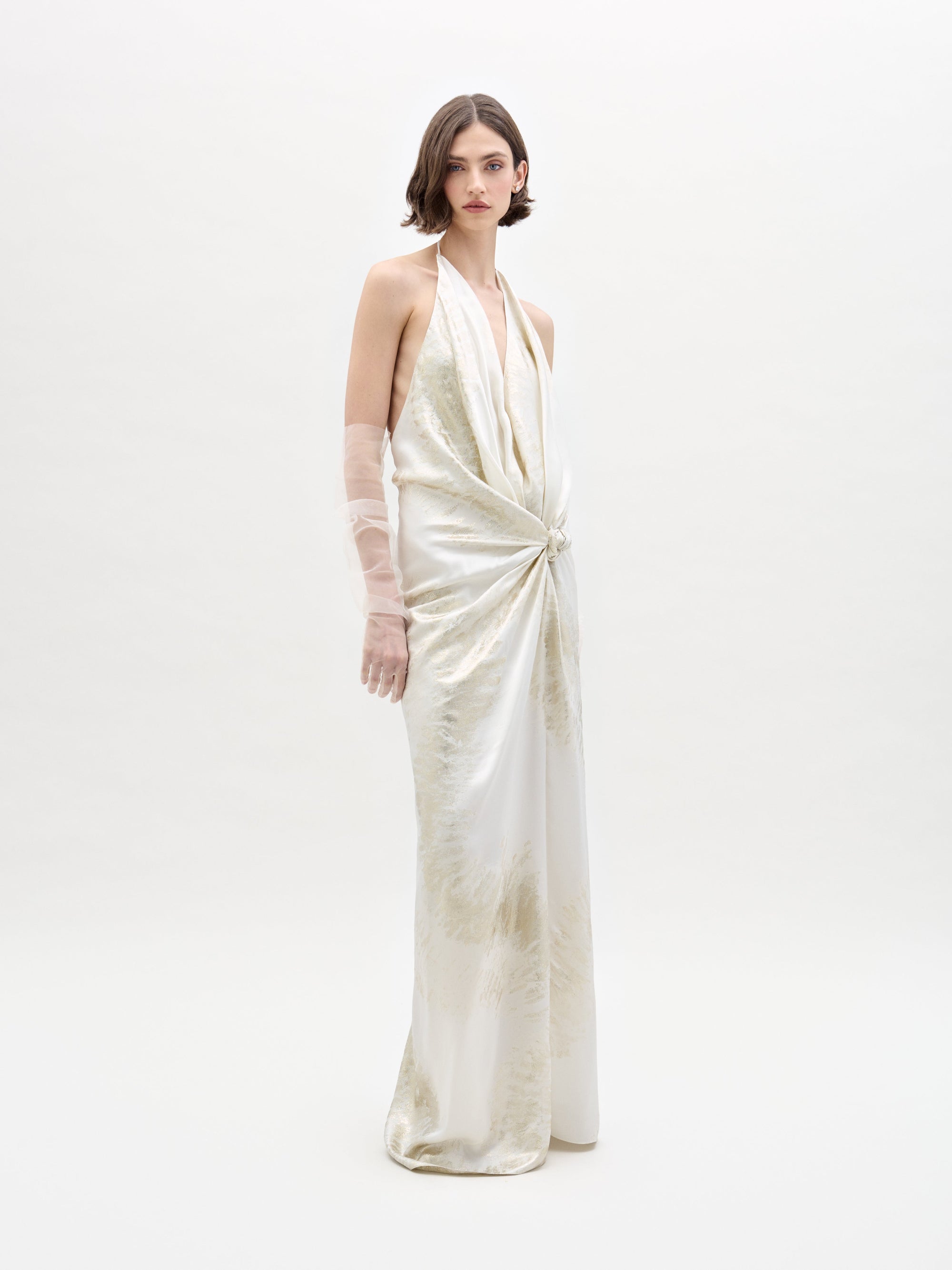 A person models the Jazmin Dress by SVT DESIGNS, featuring a twisted front halter-neck design and a subtle pattern, against a plain white background, complemented by sheer gloves.