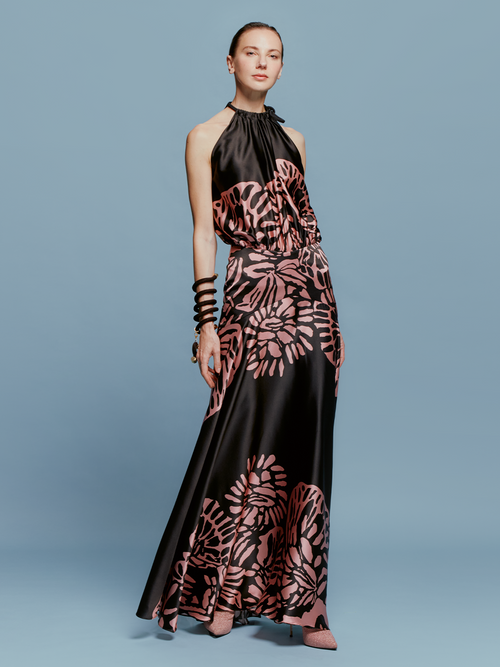 A woman is posing against a blue background, wearing the Joss Dress in Black Pink Filigrana—a black and pink floral halter neck gown—complemented by matching pink heels and black bracelets, capturing the fashion essence of September 2.