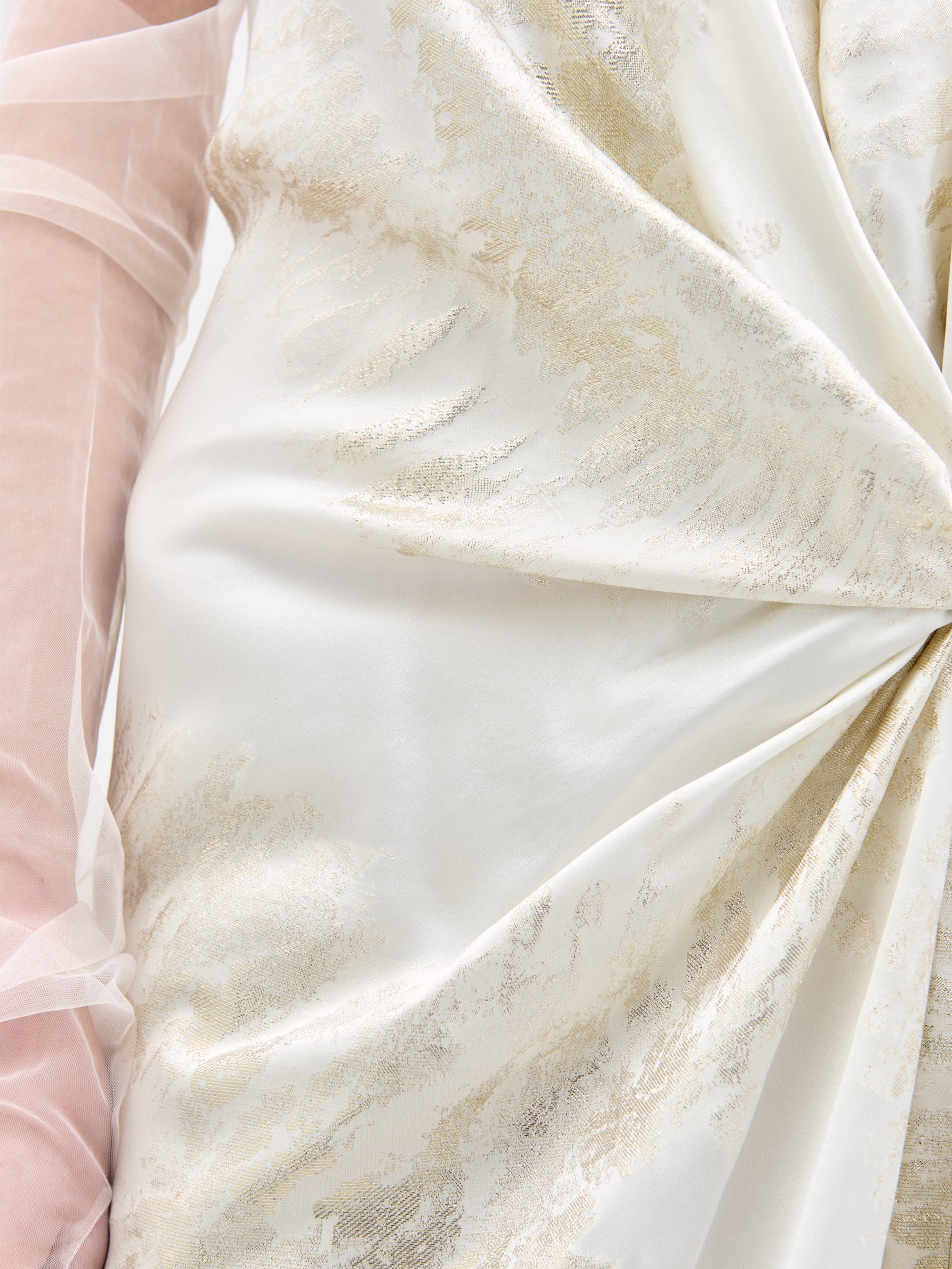 Close-up of the Jazmin Dress by SVT DESIGNS featuring white and gold swirling patterns. The fabric, tied into a knot, creates elegant folds. A translucent sleeve is partially visible on the left, suggesting a sophisticated garment design.