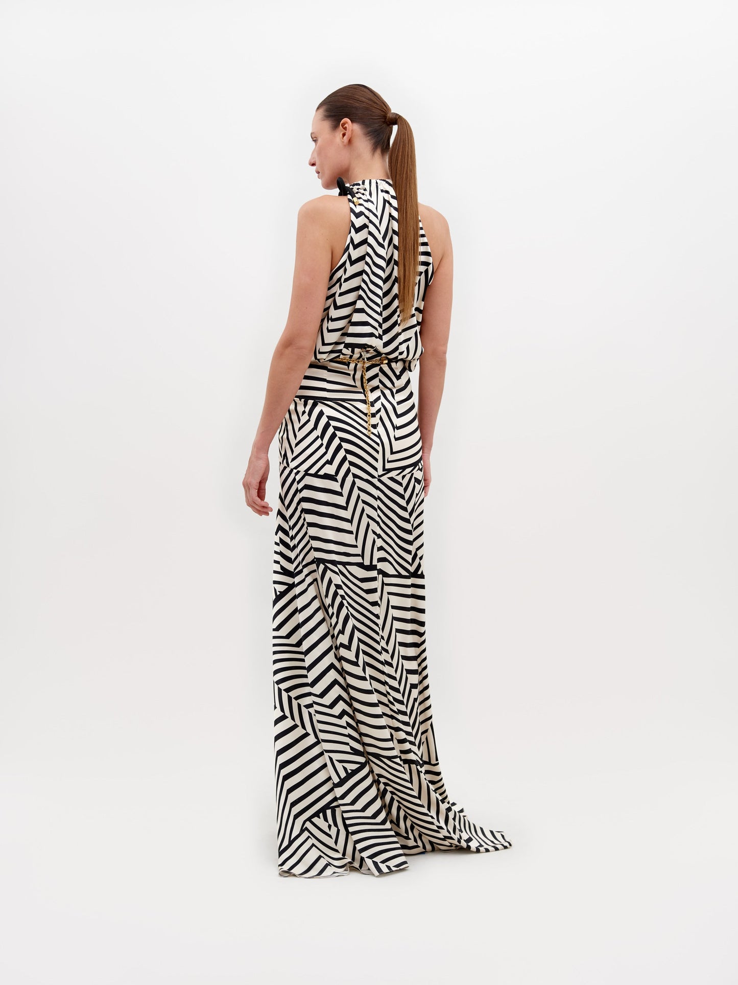 A person is wearing a Joss Dress Beige Black Stripes, accented with a black and gold belt, standing against a plain white background. This ensemble is available for pre-order now and will ship by November 15th, 2024.