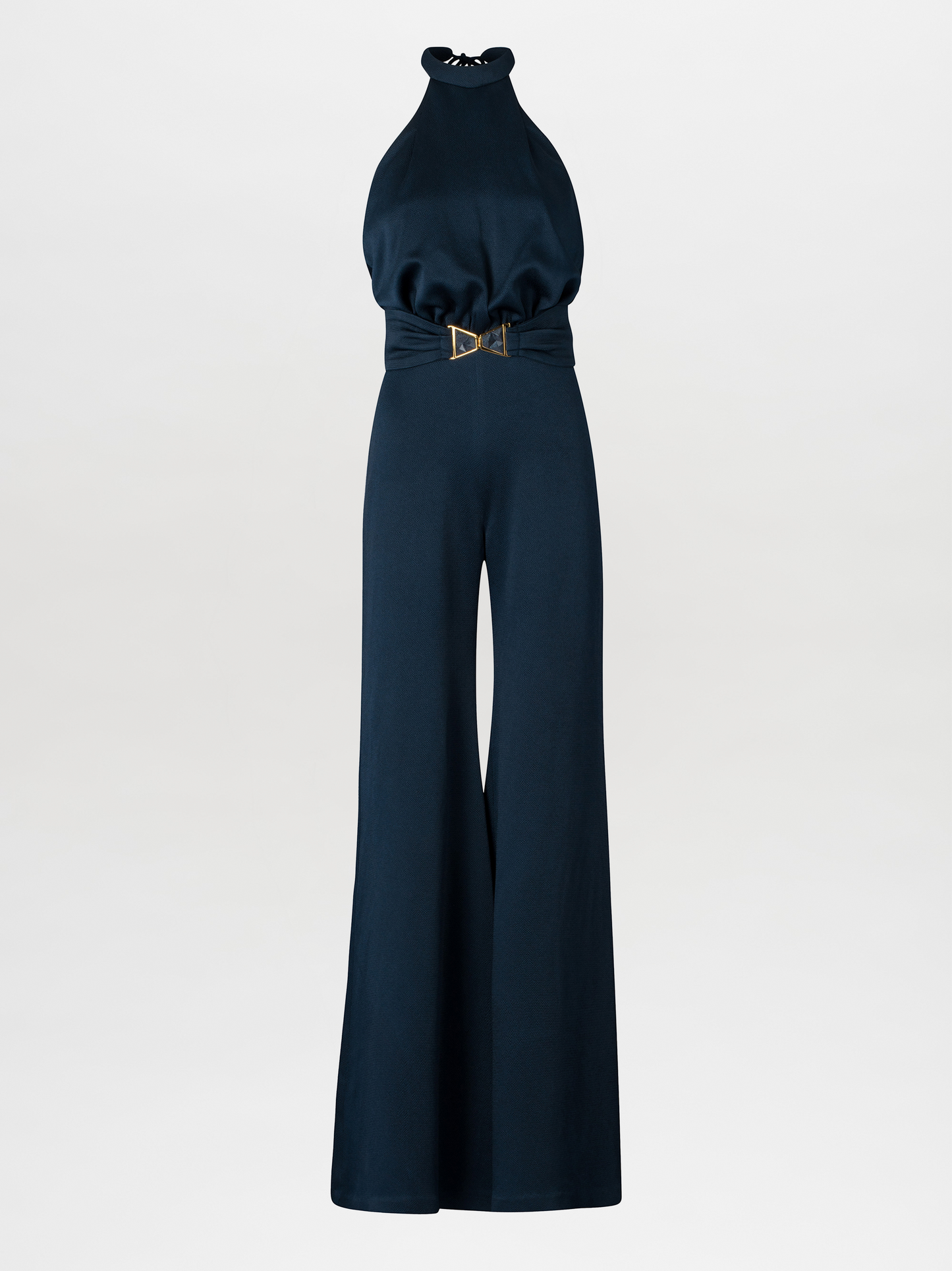 A woman wearing the Kaede Jumpsuit Navy, featuring a halter-neck design with wide-leg pants and a black belt, stands against a light blue background. She accessorizes with multiple black bracelets on one arm, exuding an air of chic nautical elegance.