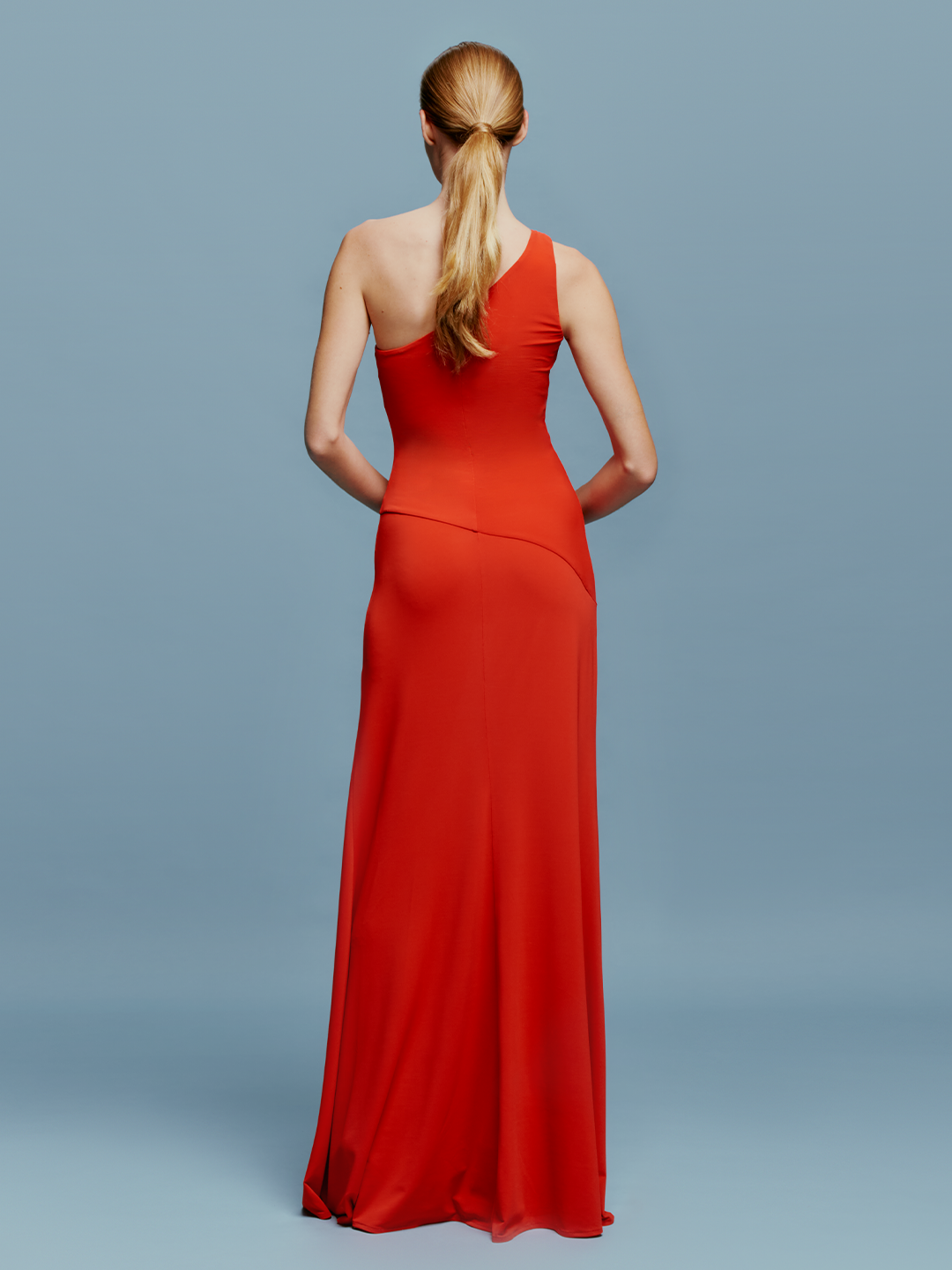 A woman in a Kanya Dress Rouge, featuring a red, one-shoulder design and floor-length cut, stands against a plain blue background. Her hair is pulled back as she calmly stands with her arms by her sides, appearing to wait for an important date such as August 19th or September 2nd.