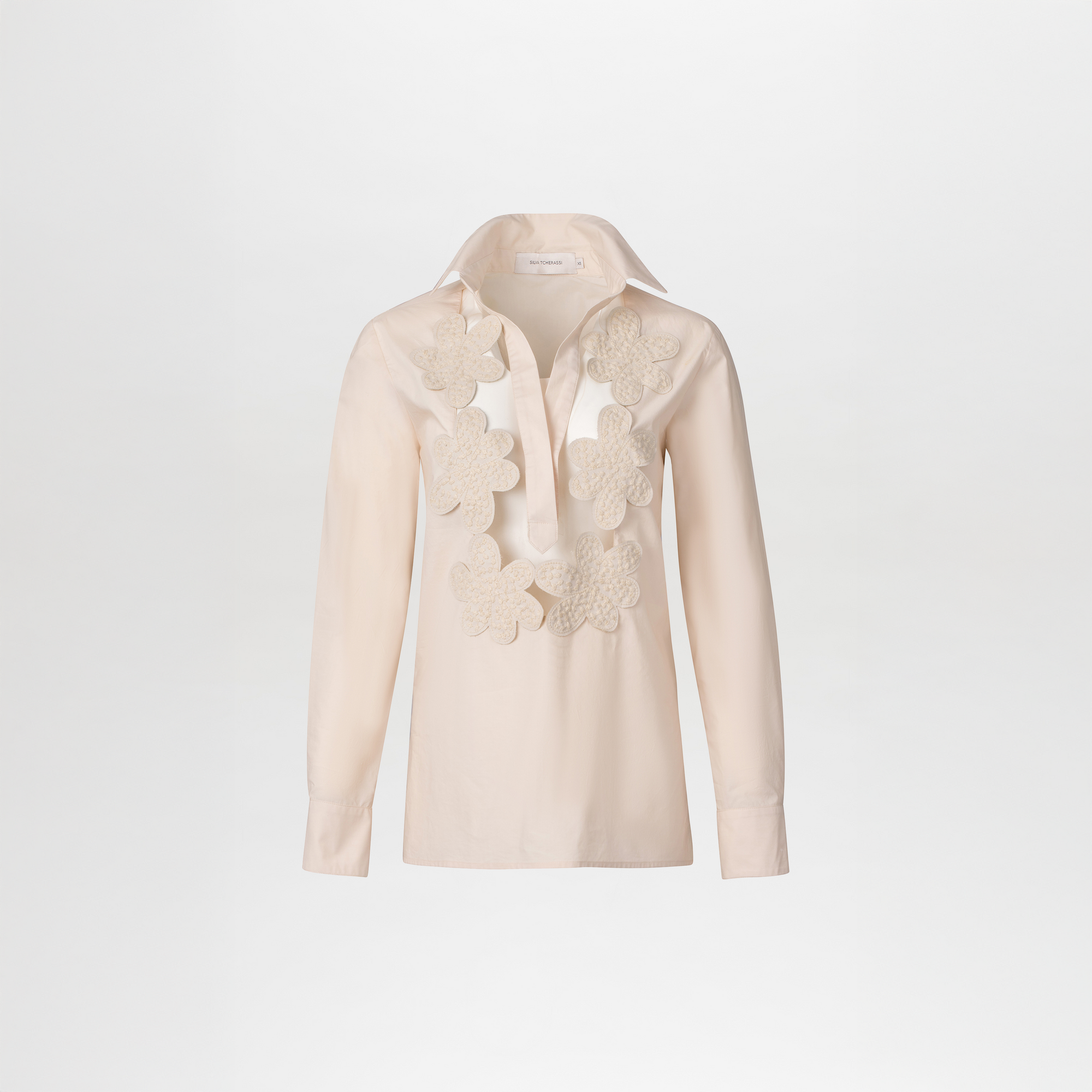 The Karole Blouse by SVT DESIGNS is a light beige, long-sleeve cotton blouse with a classic shirt collar and large textured floral lace appliqués on the front, shown against a plain white background.