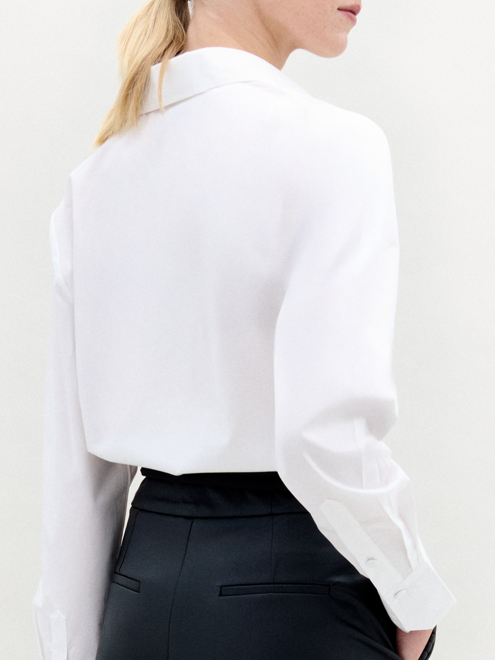 A person with blonde hair in a ponytail is shown from the back, wearing the Karole Blouse by SVT DESIGNS—a white long-sleeve blouse—paired with black high-waisted trousers, set against a plain light background.