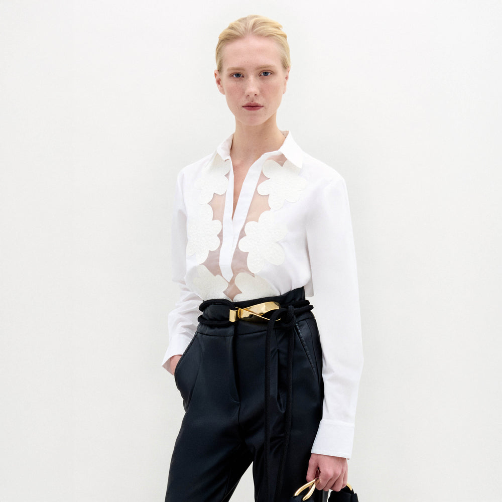 A person stands against a plain light background wearing the SVT DESIGNS Karole Blouse with large floral appliqués and sheer paneling, styled with high-waisted black pants, a gold-buckled belt, and holding a black handbag.