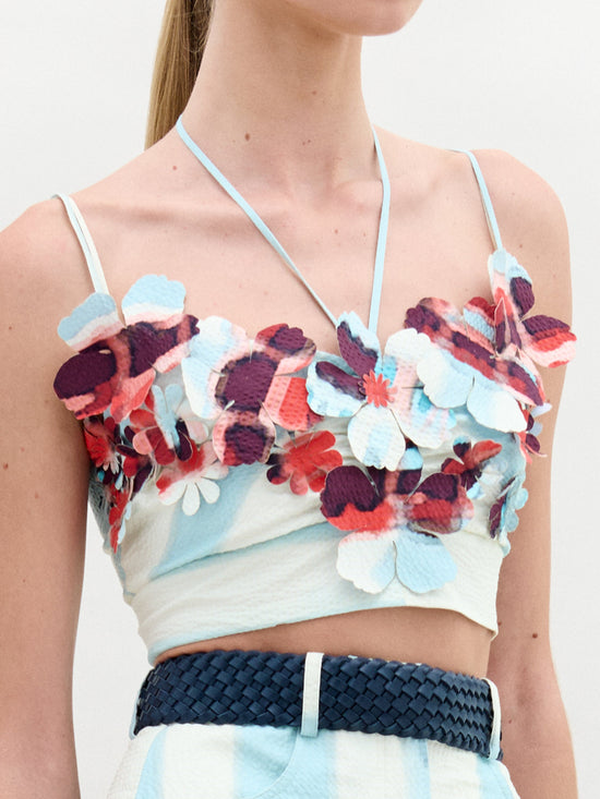 Someone wears the Kim Top by SVT DESIGNS, featuring vibrant 3D floral details with thin straps in red, blue, and white. It's paired with high-waisted pants cinched by a woven belt against a plain, light backdrop.