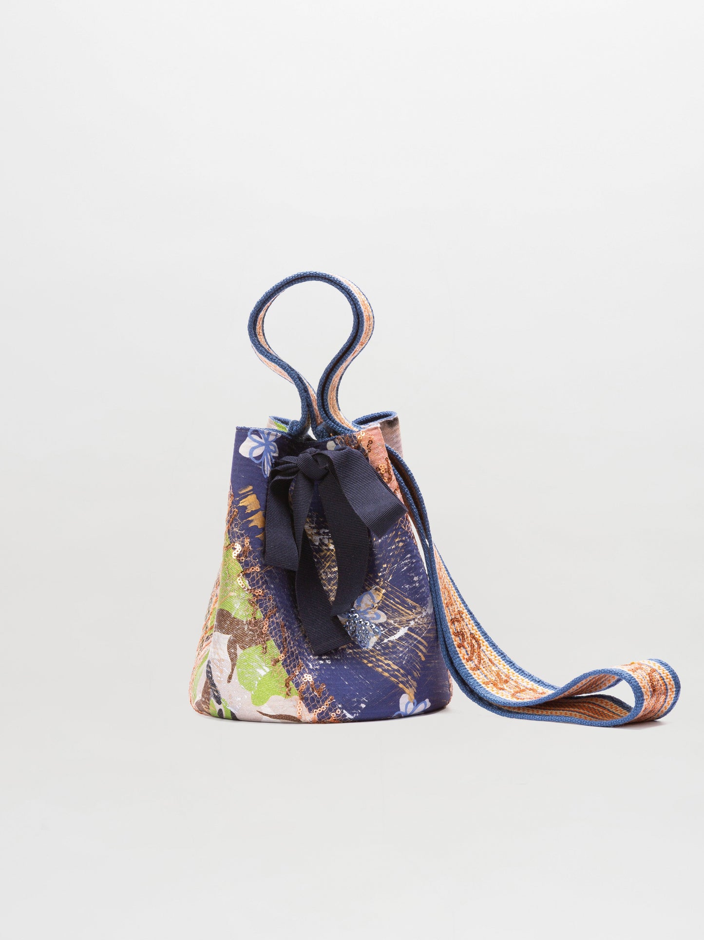 The Mochila Laaput Small by SVT DESIGNS is a compact bucket-style handbag featuring a vibrant abstract pattern, a fabric bow on the front, and a long matching strap. It stands upright against a plain light gray background.