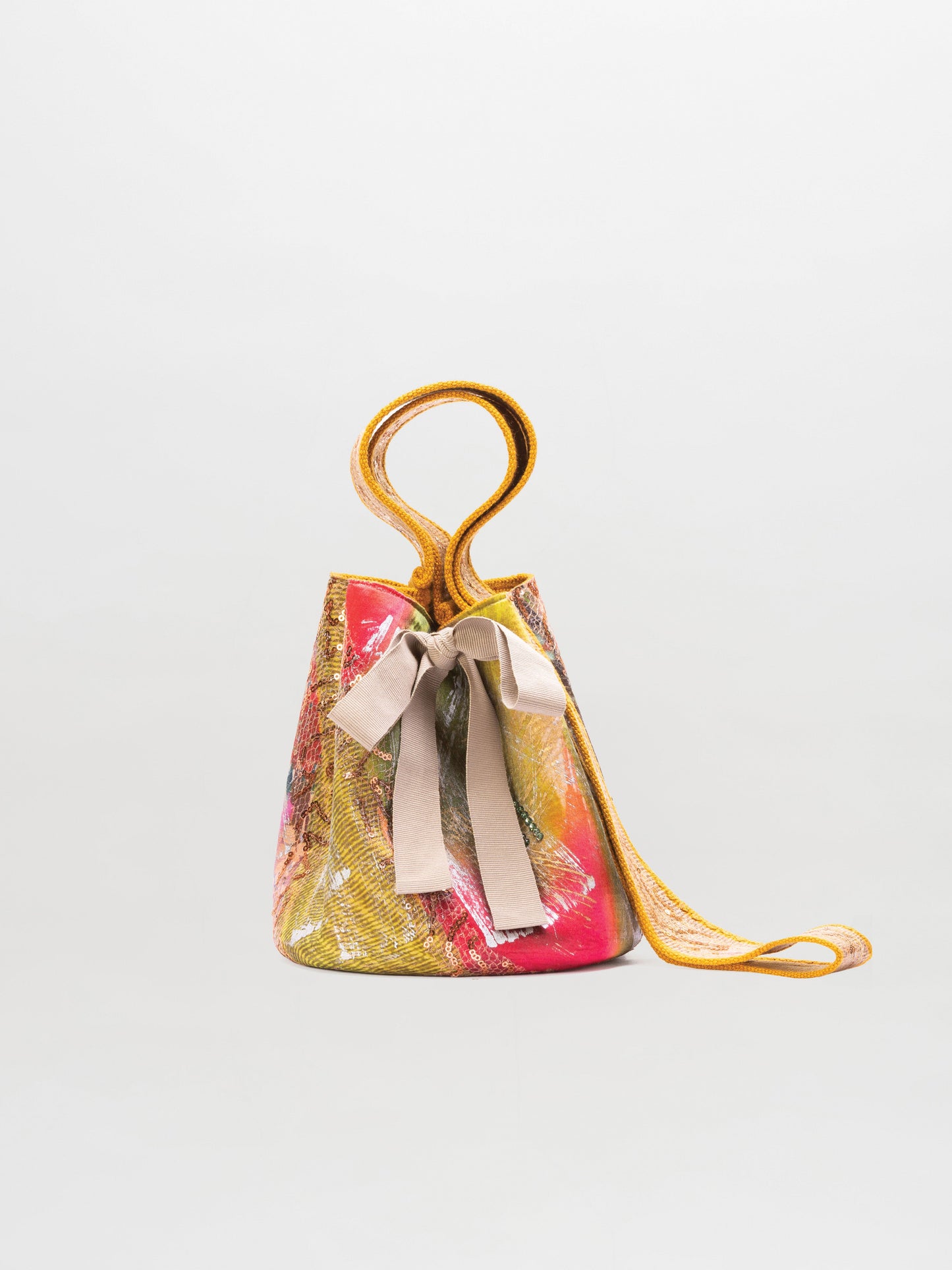 The Mochila Laaput Small by SVT DESIGNS is a small bucket-style bag featuring an abstract multicolored pattern, a yellow strap, and a beige bow at the front, shown against a plain light gray background.