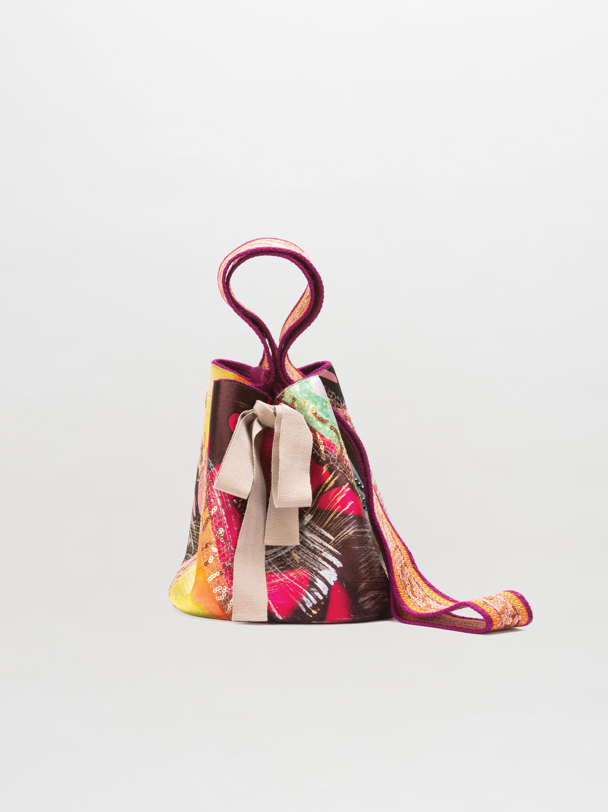 The SVT DESIGNS Mochila Laaput Small is a colorful Wayuu handbag with an abstract red, orange, and green pattern, fabric bow, and hand-woven strap, shown against a plain white background.