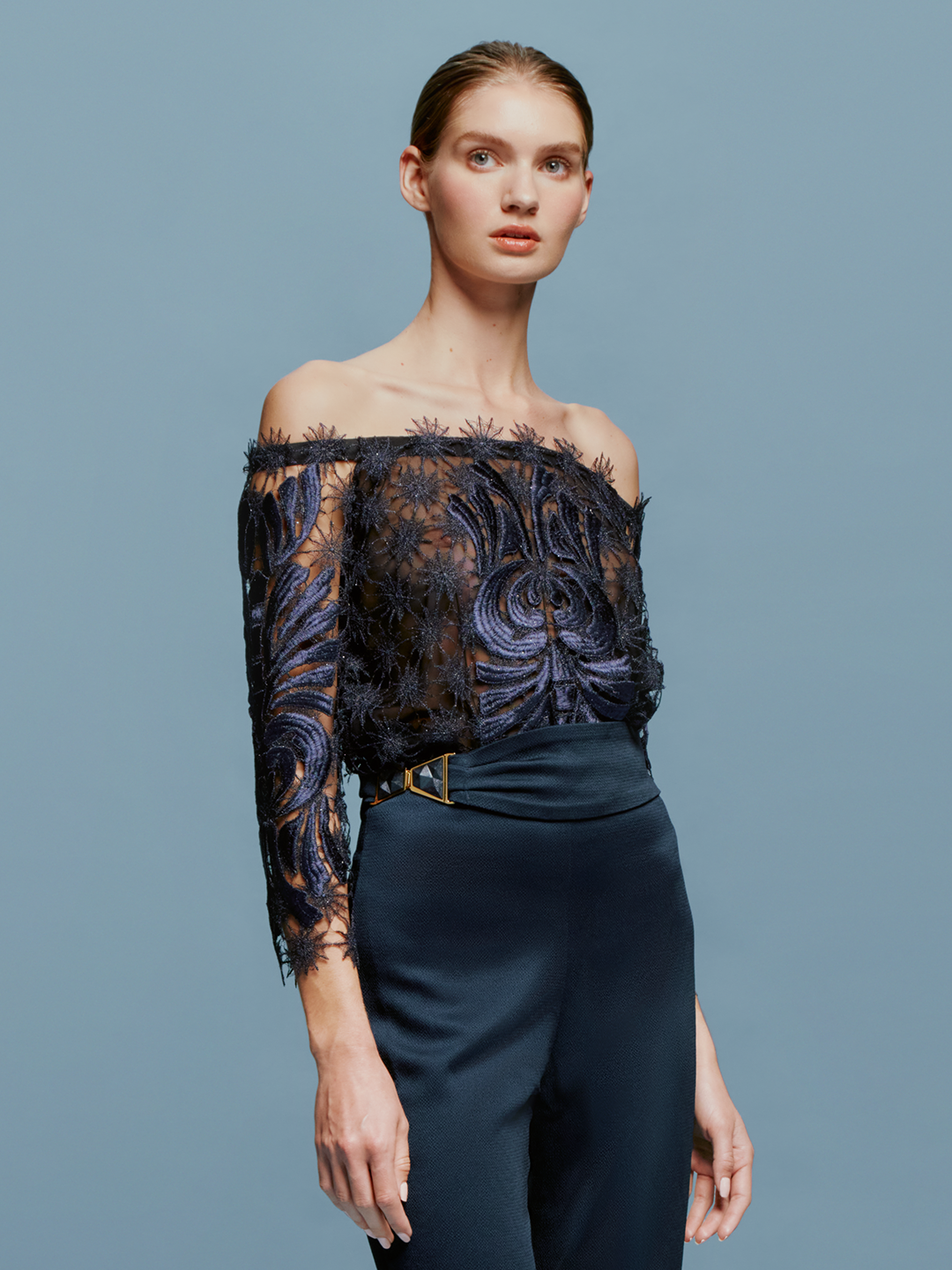 On August 19th, a person with slicked-back hair wears the Lala Blouse Navy with intricate patterns and high-waisted navy pants against a plain blue background.