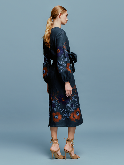 A person stands against a blue background wearing the Laoise Dress in Navy Ochre Flower Embroidery, paired with beige high-heeled sandals, perfect for an August 19th or September 2nd outing.