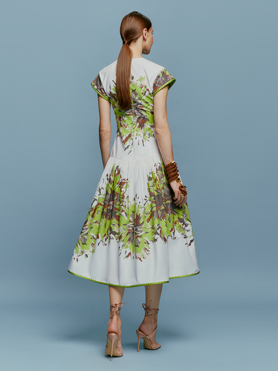 A woman in the Lara Dress Floral Camo, featuring a white base with green floral patterns, stands against a blue background. She is wearing beige high-heeled shoes and wooden bangles, perfectly capturing the essence of August 19th.