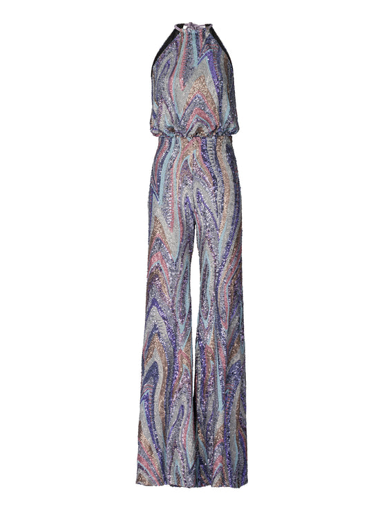 Against a plain blue background, a person stands poised in the Lauretta Jumpsuit Sequin Waves, featuring a sleeveless, high-neck design with a colorful, swirling pattern. The outfit showcases wide-leg pants and comes with a belt—perfectly capturing the style for upcoming events between August 19th and September 2nd, 2024.