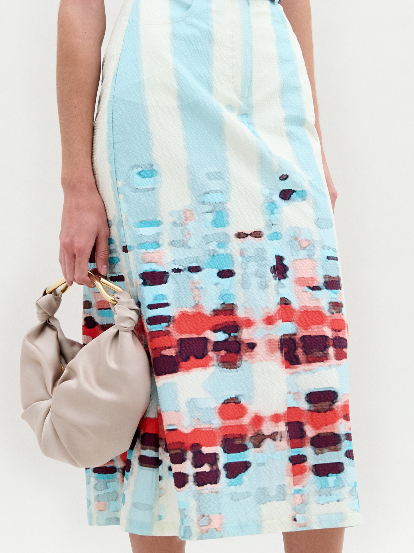 A person holding a beige bag with ruched details and a circular handle is wearing the Layan Skirt, a pastel blue midi skirt by SVT DESIGNS, featuring abstract red and pink patterns with vertical stripes. The fitted silhouette stands out against the plain background, highlighting this unique ensemble.