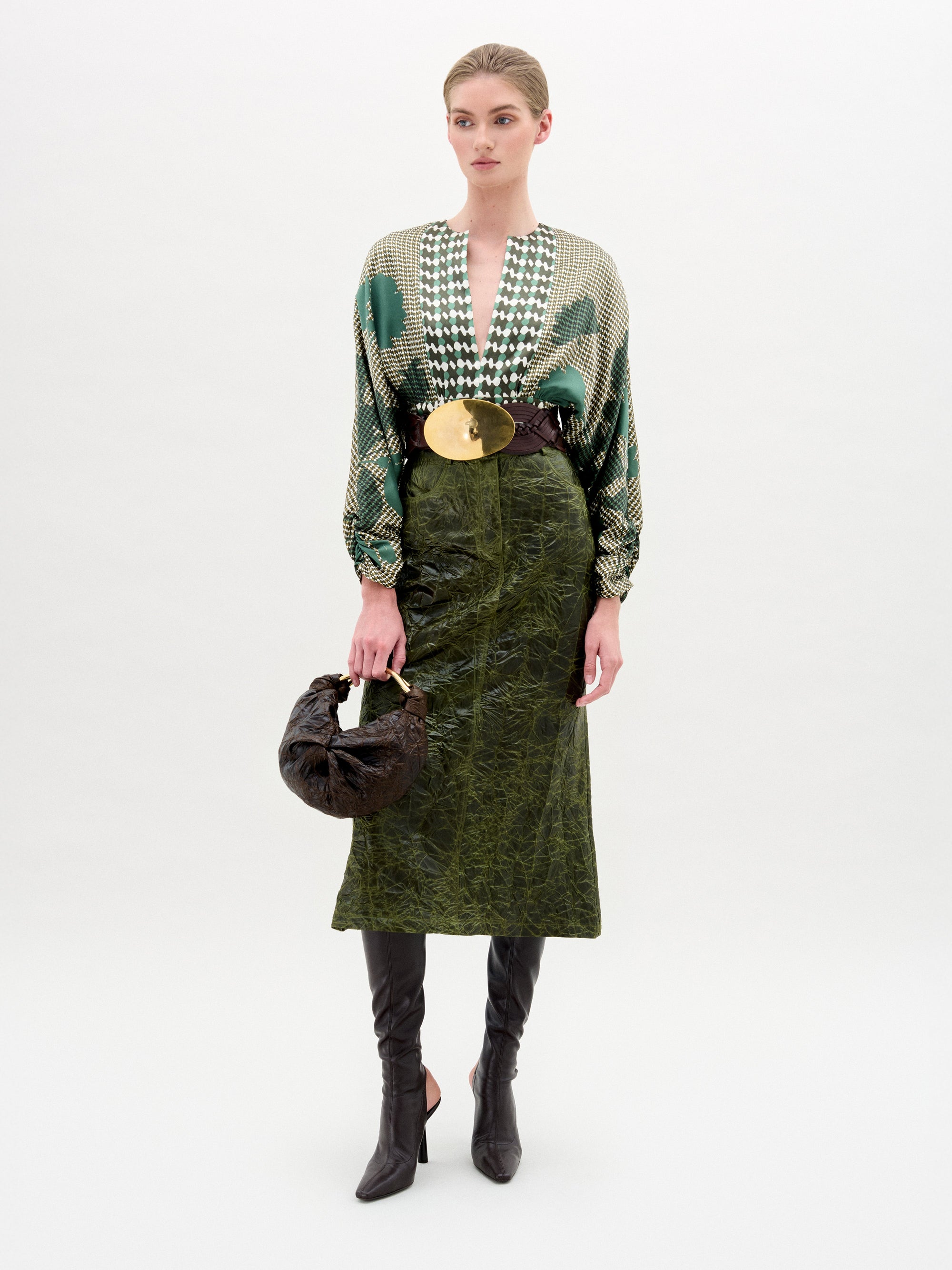 A woman stands against a plain background wearing a patterned green blouse, a wide belt with a large gold buckle, SVT DESIGNS' Layan Skirt made in Colombia, black knee-high boots, and holding a brown handbag.